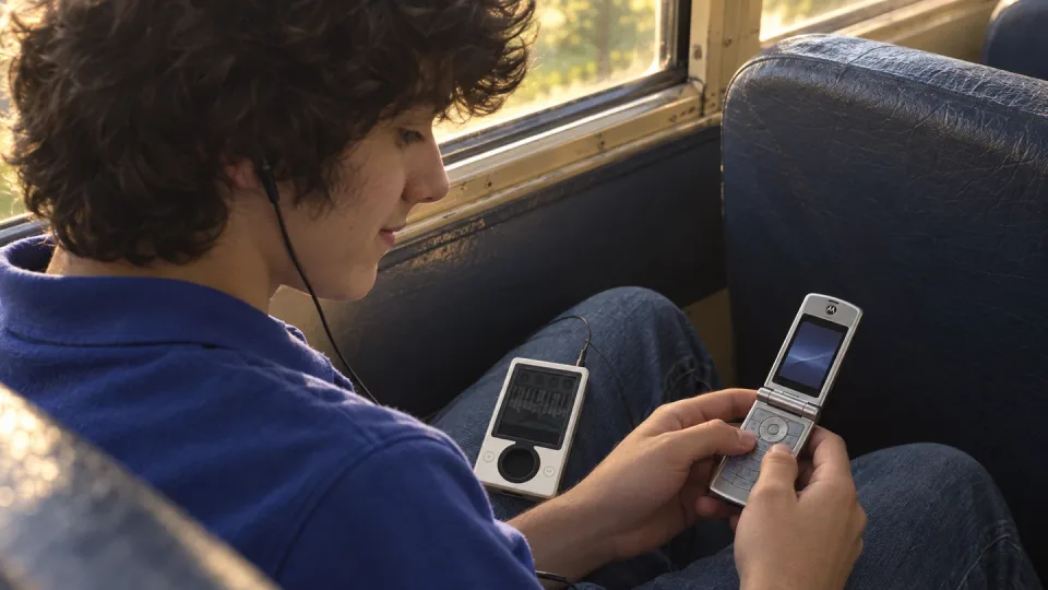 AI reconstruction of teenage me on the bus, with a white Zune on my lap and a silver flip phone in my hands